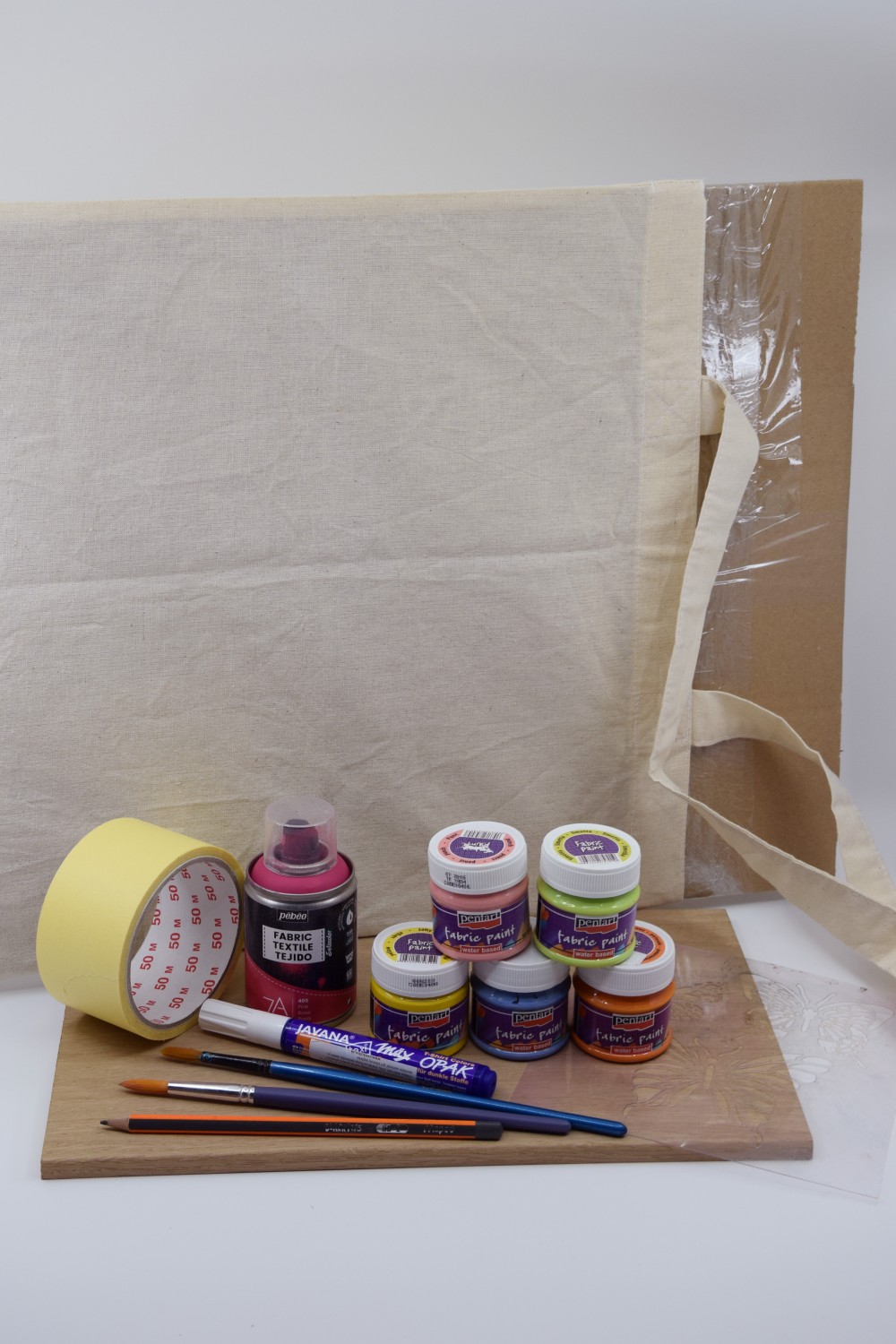 Na obrázku sú farby na textil, s fixkami, štetcami a ceruzkou postavené pred plátennou taškou s kartónom vo vnútri.